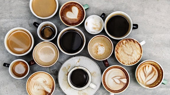 15 cups of coffee in different cups on different saucers, some with foam some without. Each one with foam has a different design or pattern drawn in the foam