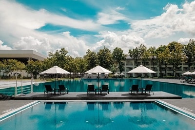 Landing page of a website for a fictional hotel. The image used was of the pool area, in front is a kiddy pool, past that are three umbrellas each with two lounging chairs, followed by the main pool with more umbrellas and chairs. The background is trees and hotel rooms under a blue sky with pillowy and wispy clouds.